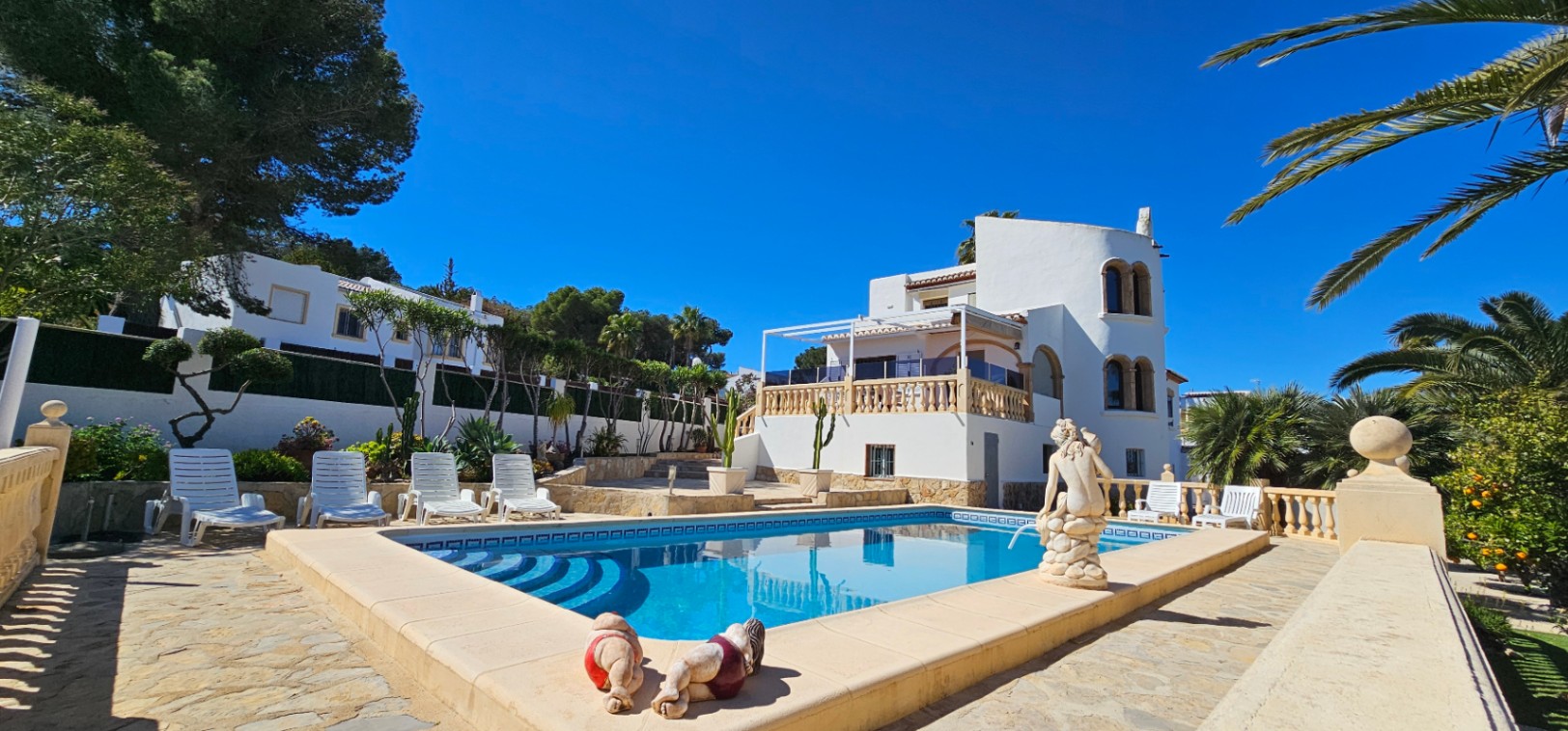 Piscina rectangular con escaleras romanas rodeada de una amplia terraza de piedra, tumbonas blancas y una villa de estilo mediterráneo con torre circular.