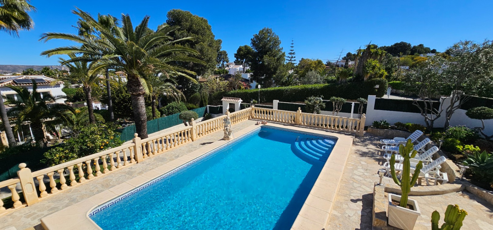 Piscina rectangular con escaleras romanas rodeada por una amplia terraza de piedra, balaustrada clásica y un jardín mediterráneo con palmeras maduras.