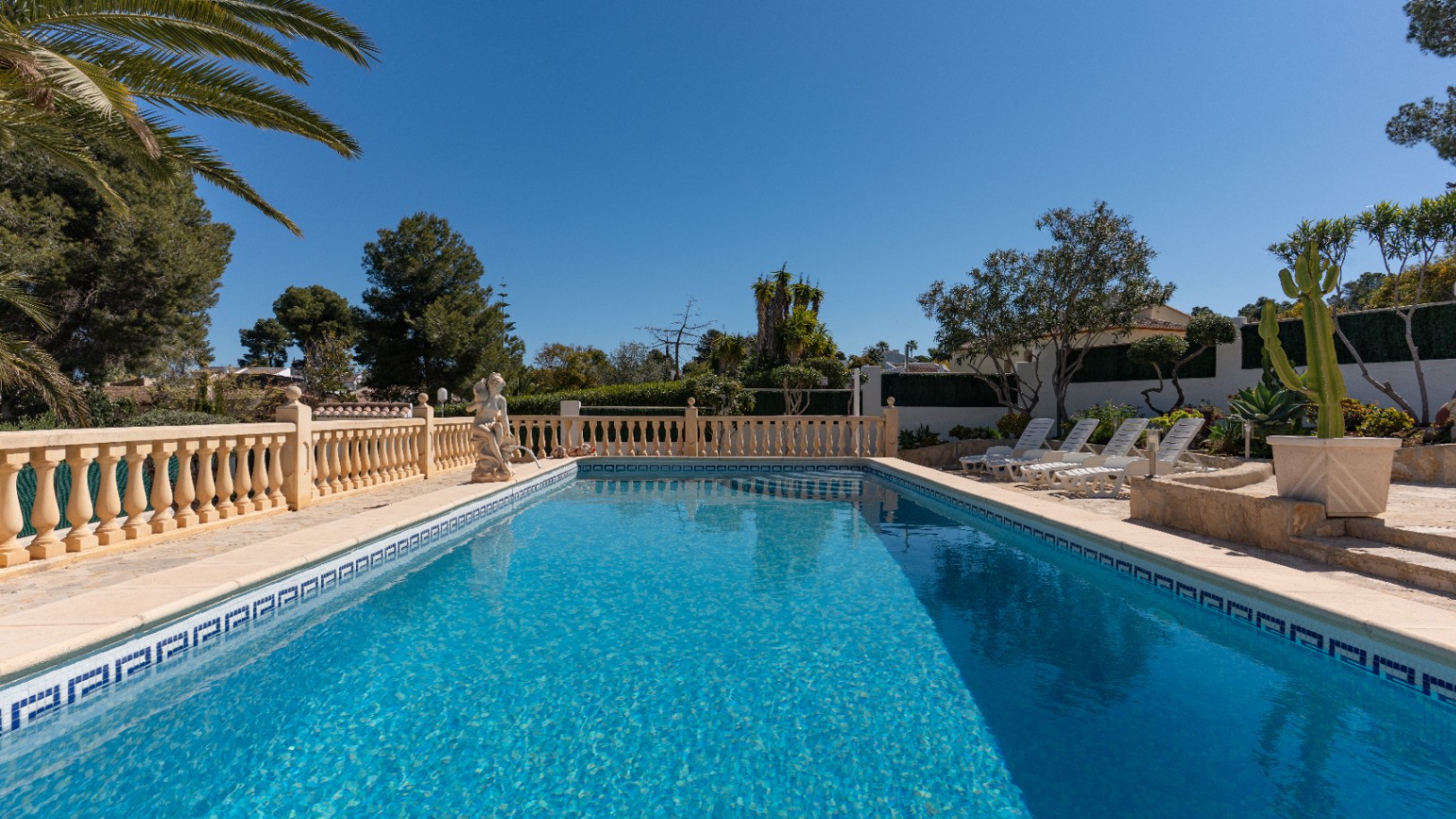 Piscina rectangular con revestimiento de mosaico azul, rodeada por una terraza de piedra con balaustrada clásica y zona de solárium con tumbonas bajo el sol mediterráneo.
