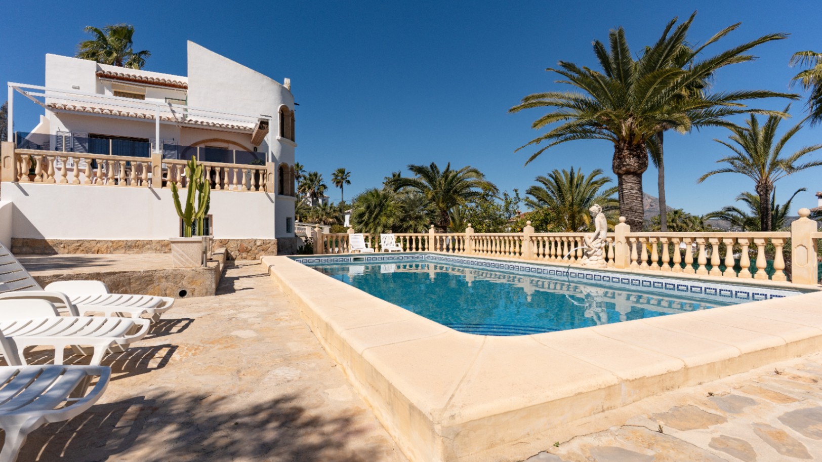 Piscina rectangular rodeada de una amplia terraza de piedra con tumbonas, palmeras maduras y fachada blanca con balaustradas tradicionales en Jávea.