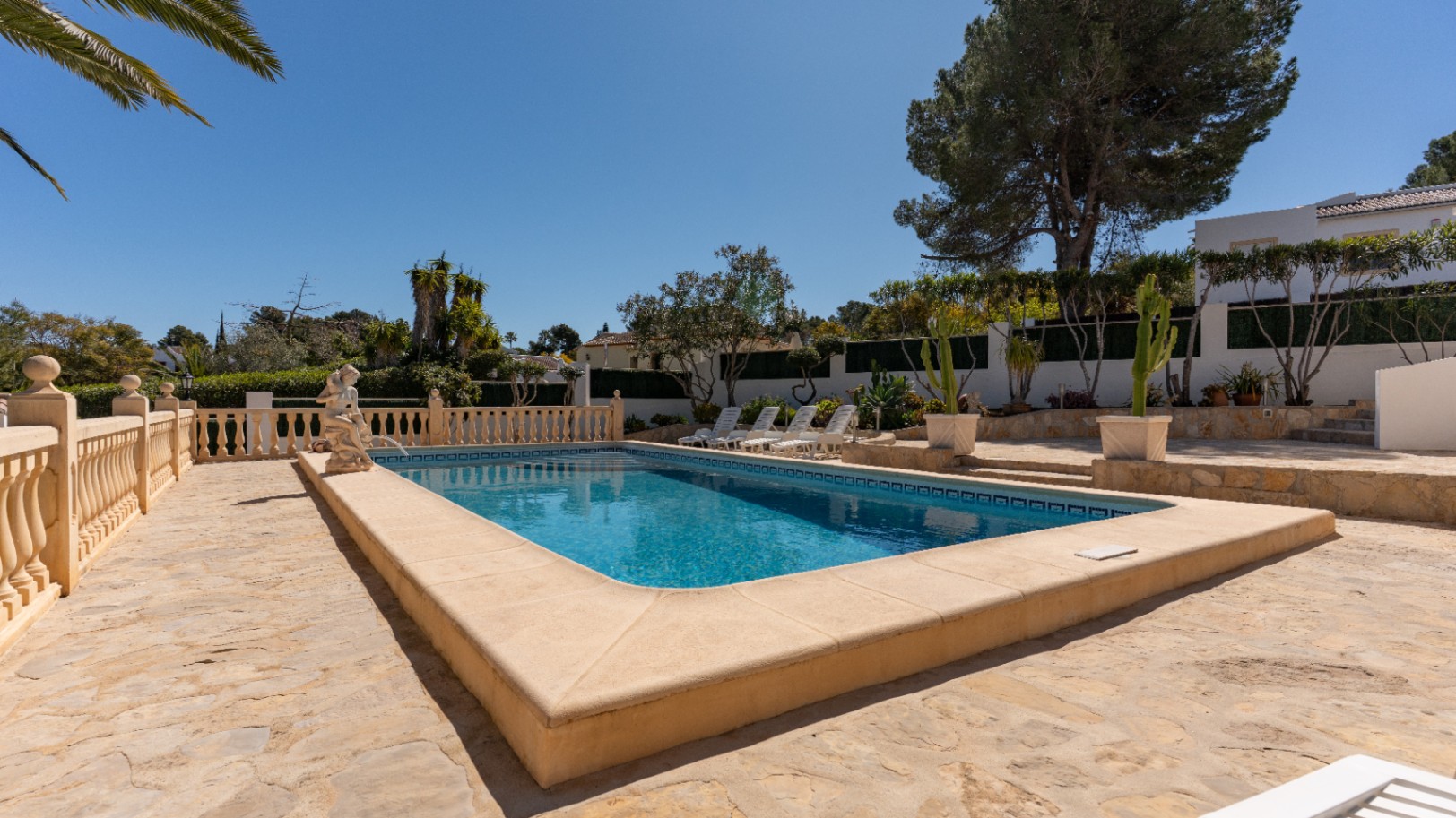 Piscina rectangular rodeada de una amplia terraza de piedra natural, con balaustrada clásica, estatua decorativa y zona de solárium junto a vegetación mediterránea.