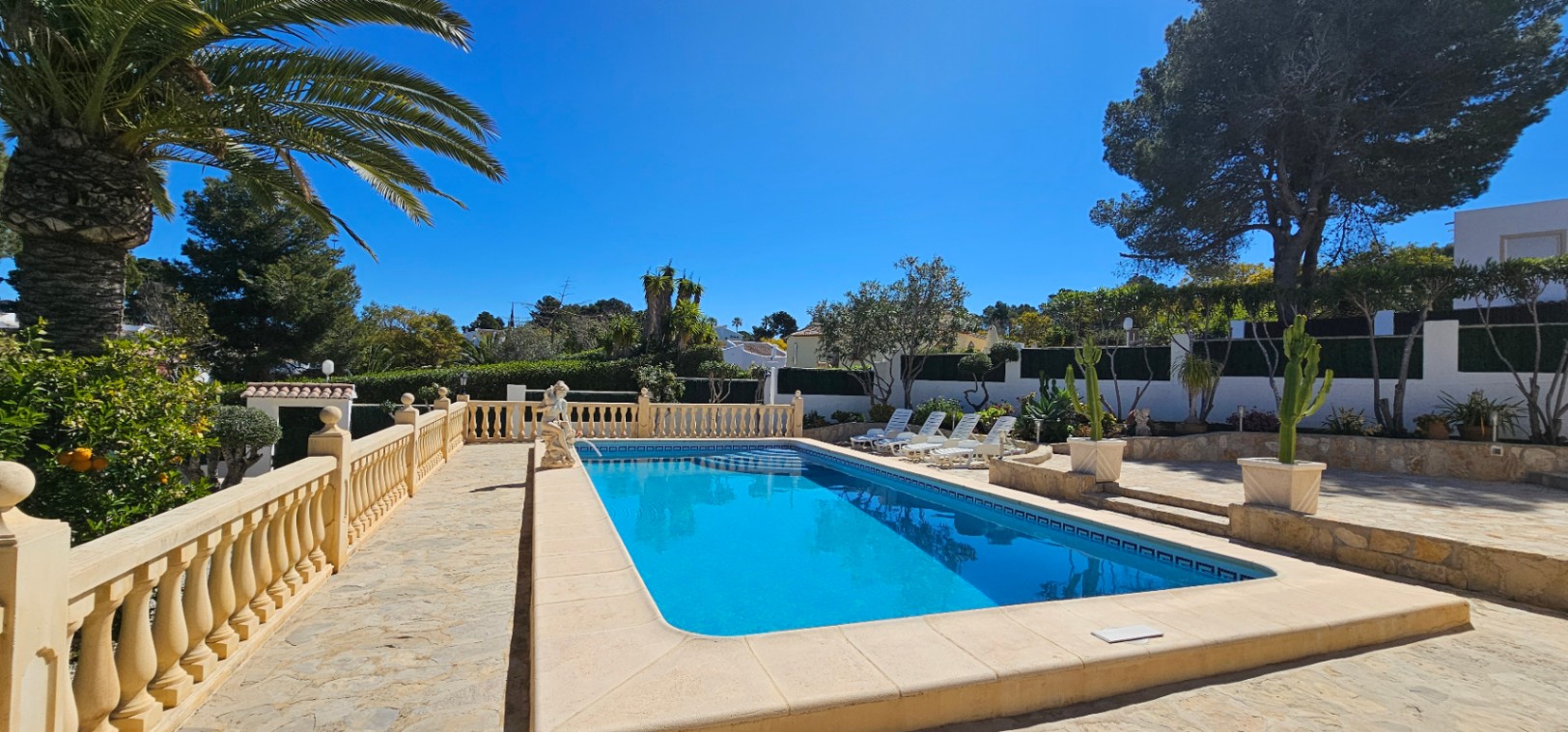 Piscina rectangular rodeada de una amplia terraza de piedra natural con balaustrada clásica, zona de tumbonas y vegetación mediterránea bajo un cielo despejado.