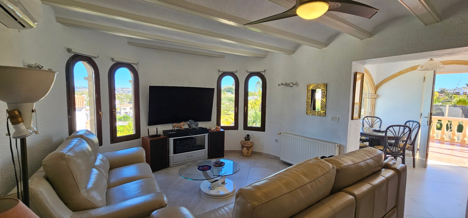 Salón con sofás de cuero, techo de vigas blancas y ventanas de arco. La estancia integra una zona de televisión y conecta con el comedor mediante un arco tradicional.