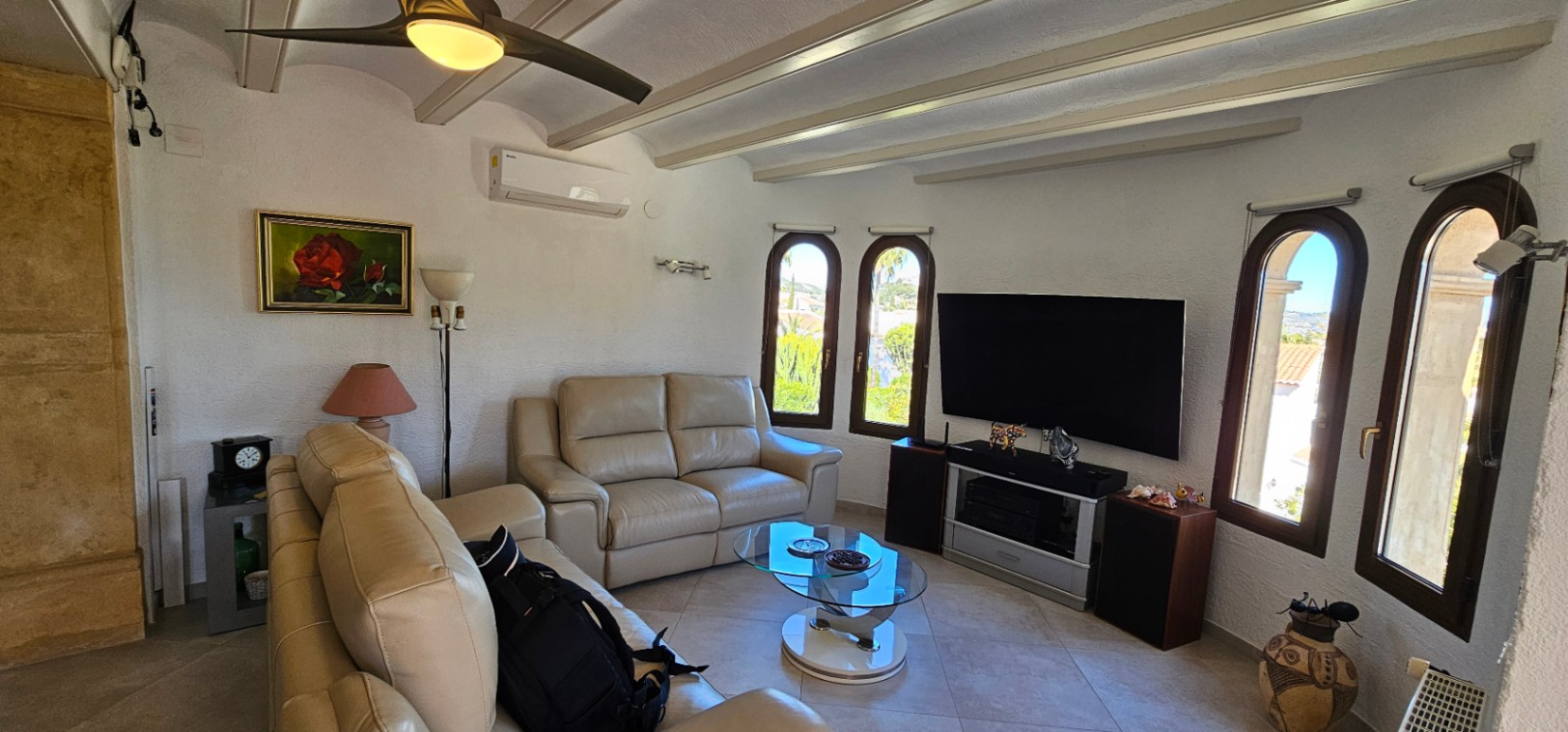 Salón luminoso con sofás de cuero beige, techos con vigas expuestas, aire acondicionado y ventanas de arco que permiten la entrada de abundante luz natural.