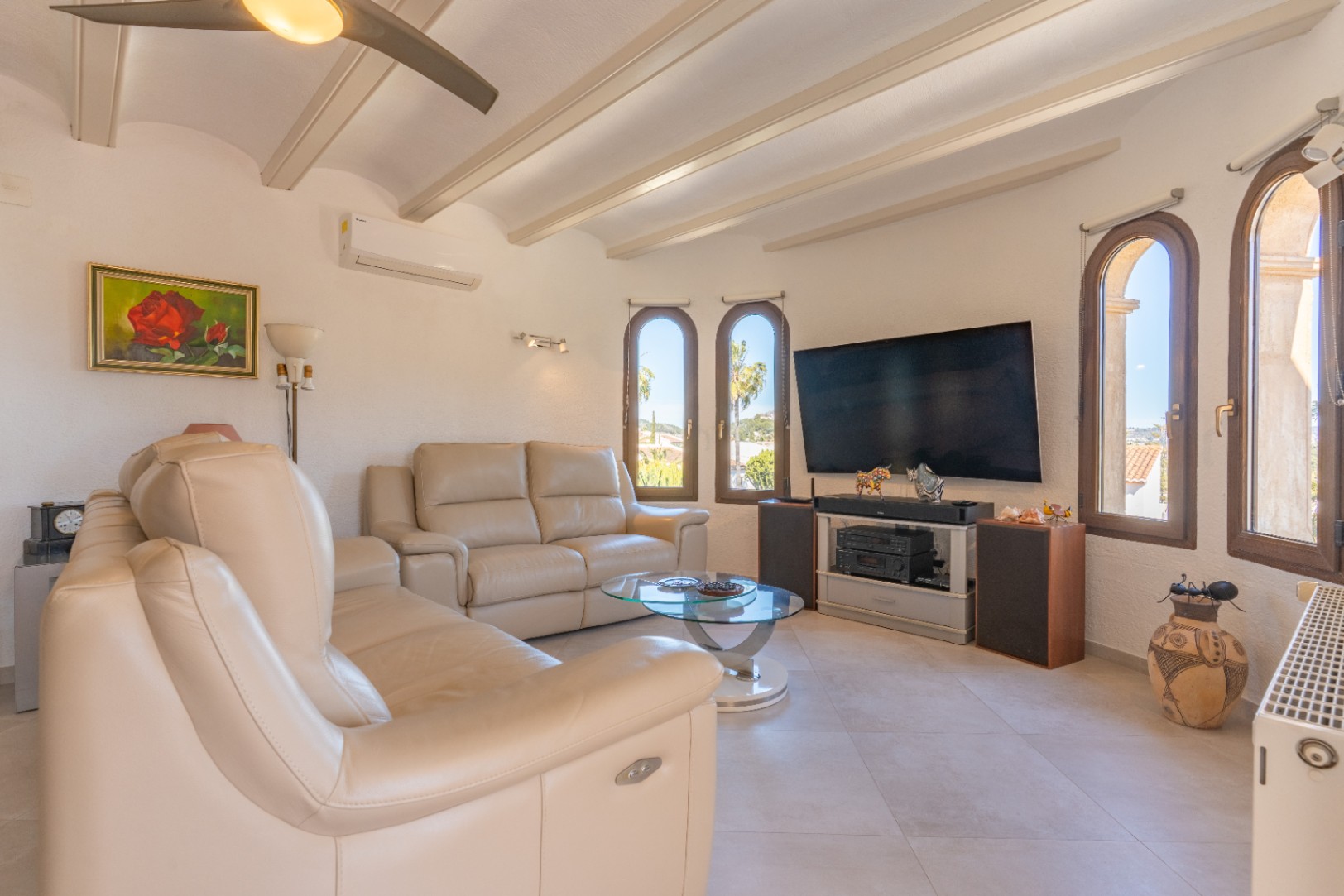 Salón luminoso con vigas vistas, sofás de cuero crema y ventanas de arco. Cuenta con aire acondicionado, ventilador de techo y un sistema de entretenimiento completo.