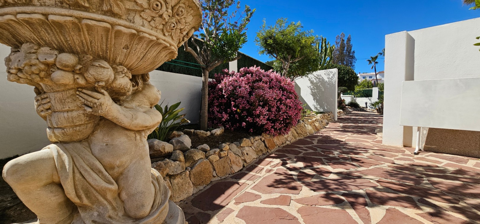 Sendero de piedra irregular flanqueado por una estatua ornamental y vegetación mediterránea, incluyendo adelfas rosadas bajo un cielo despejado.