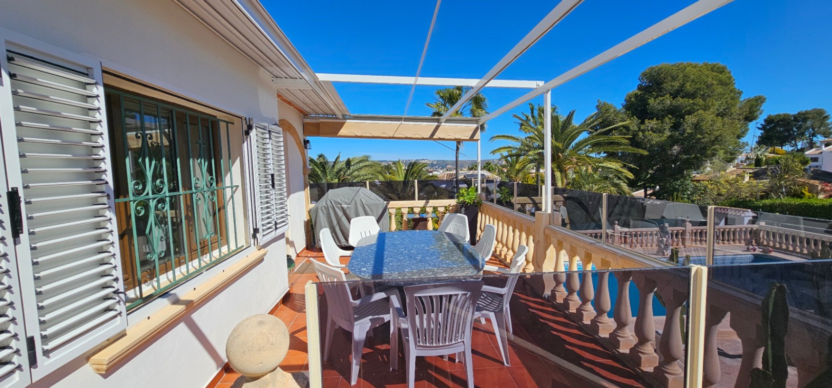 Terraza elevada con suelo de terracota, mesa de granito y cerramiento de cristal. Ofrece vistas despejadas a la piscina y al entorno mediterráneo bajo un toldo retráctil.