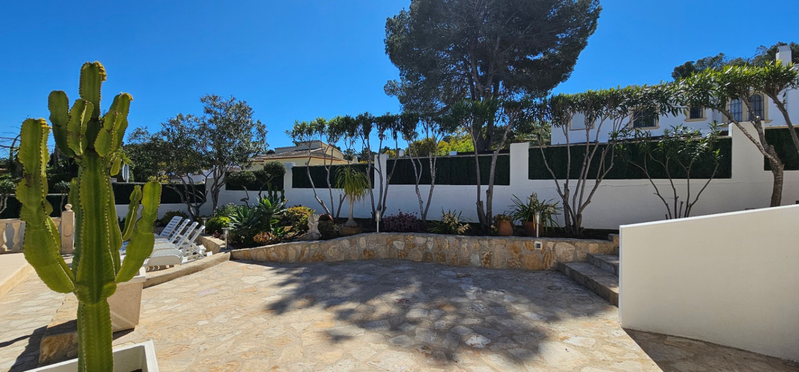 Terraza exterior con pavimento de piedra natural y muros de contención. El jardín incluye cactus y vegetación mediterránea junto a una zona de descanso.