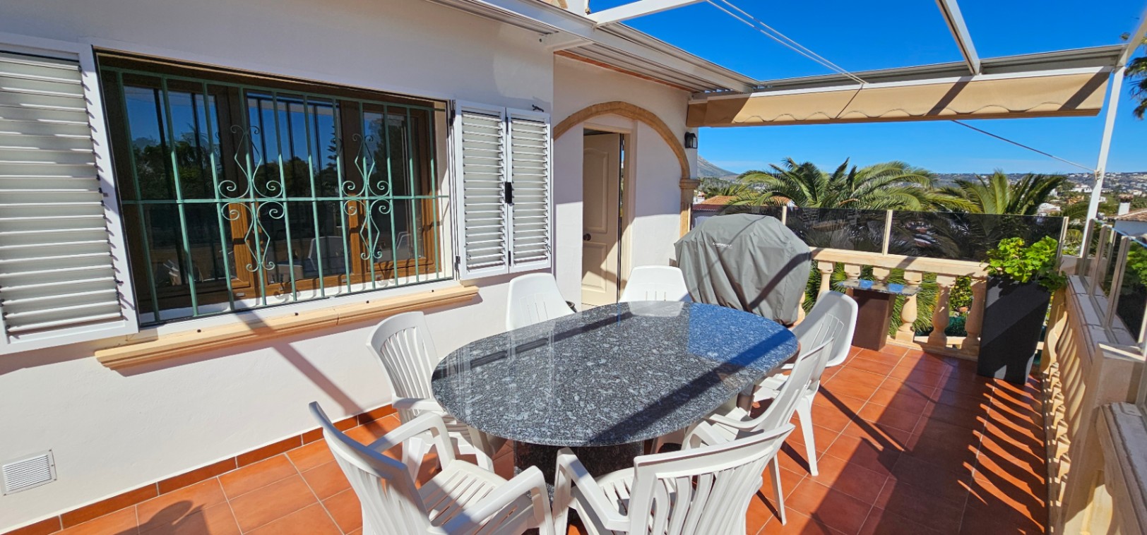 Terraza exterior con suelo de barro, mesa de granito y toldo retráctil. Destaca la balaustrada de piedra y las ventanas con rejas decorativas y contraventanas blancas.