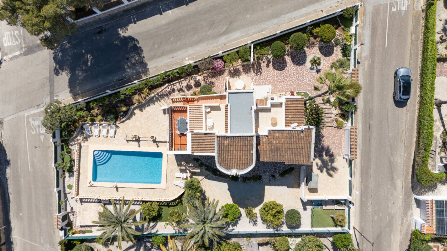 Vista aérea cenital de la propiedad en parcela de esquina, destacando la piscina rectangular, las terrazas pavimentadas y el jardín mediterráneo con palmeras.