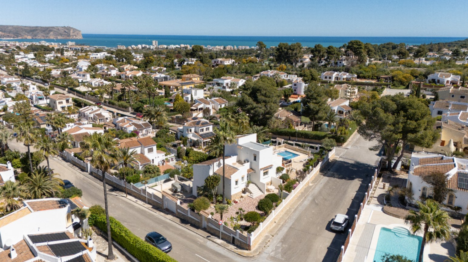 Vista aérea de la villa blanca situada en una parcela de esquina en Pinosol, Jávea. Destaca su piscina privada, jardín con palmeras y vistas despejadas hacia el mar Mediterráneo.