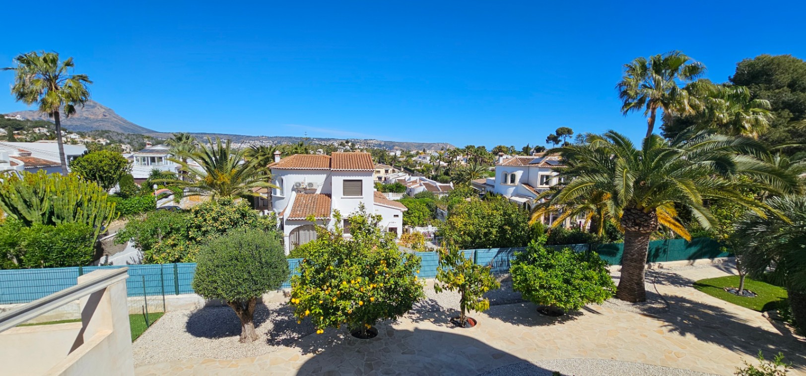 Vista panorámica desde la terraza superior que muestra el jardín con árboles frutales, palmeras y vistas despejadas hacia el Montgó y el entorno residencial de Jávea.
