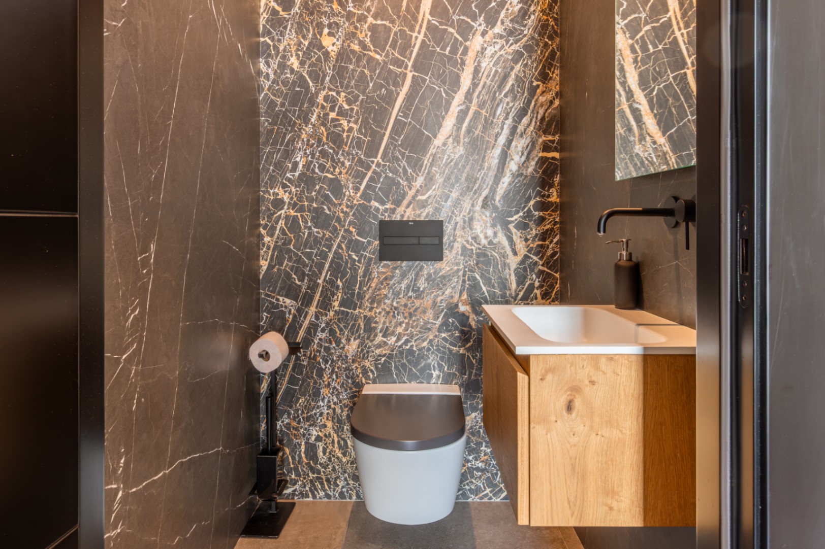 Aseo de invitados con pared de mármol oscuro y vetas doradas, inodoro suspendido y mueble de lavabo en madera de roble con grifería negra mate.