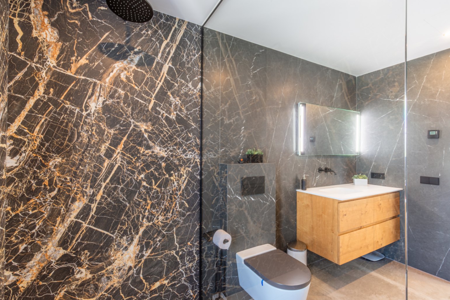 Baño moderno con ducha de efecto lluvia, revestimiento de porcelánico oscuro veteado, mueble de lavabo suspendido en madera y espejo retroiluminado.