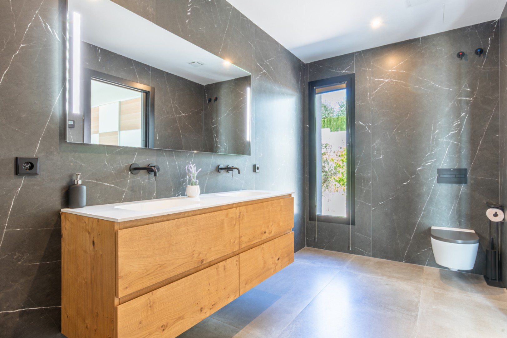 Baño moderno con revestimiento de mármol oscuro, mueble de madera de doble seno y grifería negra mate. Incluye inodoro suspendido y un gran espejo con iluminación LED integrada.