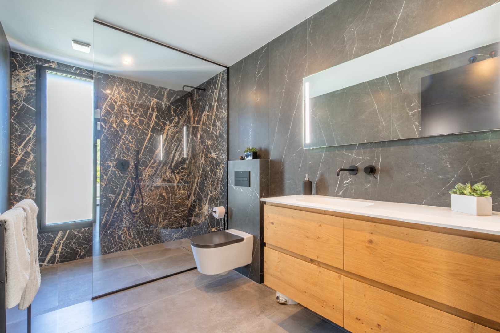 Baño moderno con revestimiento porcelánico efecto mármol oscuro, mueble de madera natural con lavabo integrado y amplia ducha italiana con mampara de cristal.