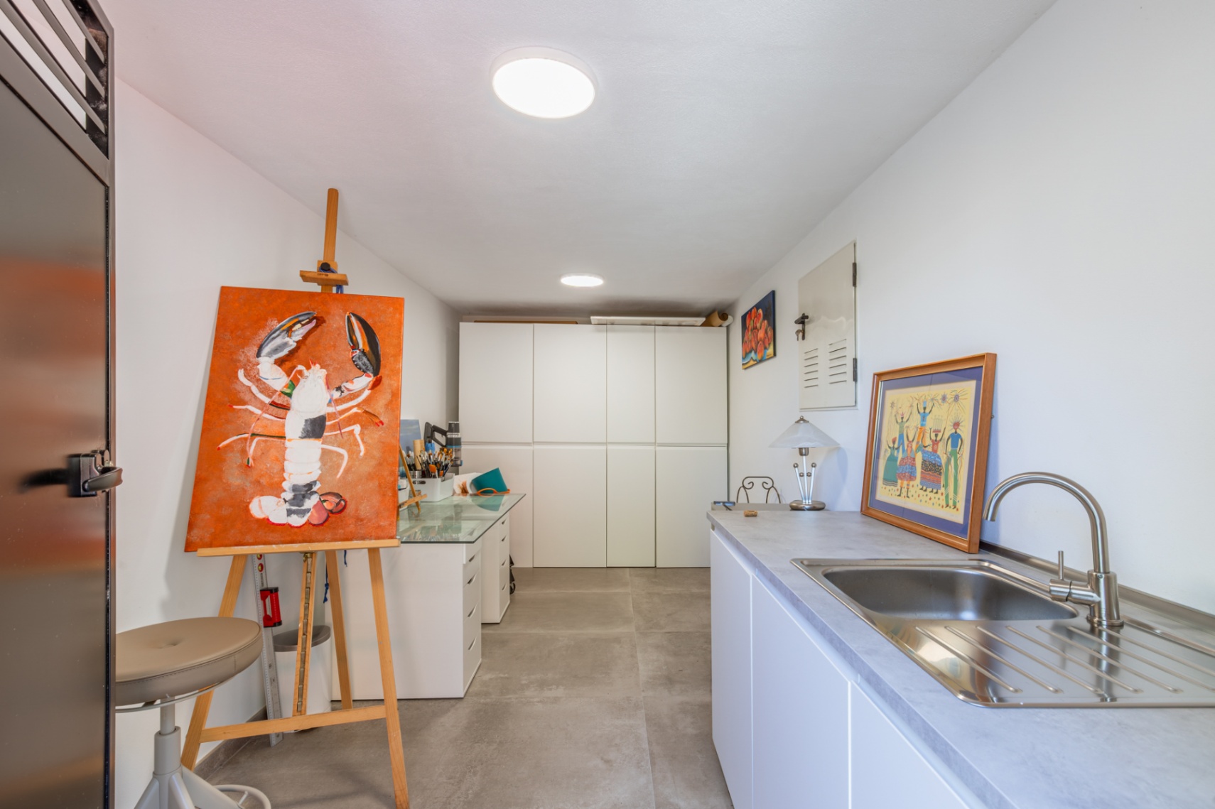 Espacio versátil de estudio o lavadero con armarios empotrados blancos, fregadero de acero inoxidable y zona de trabajo sobre suelo porcelánico gris.