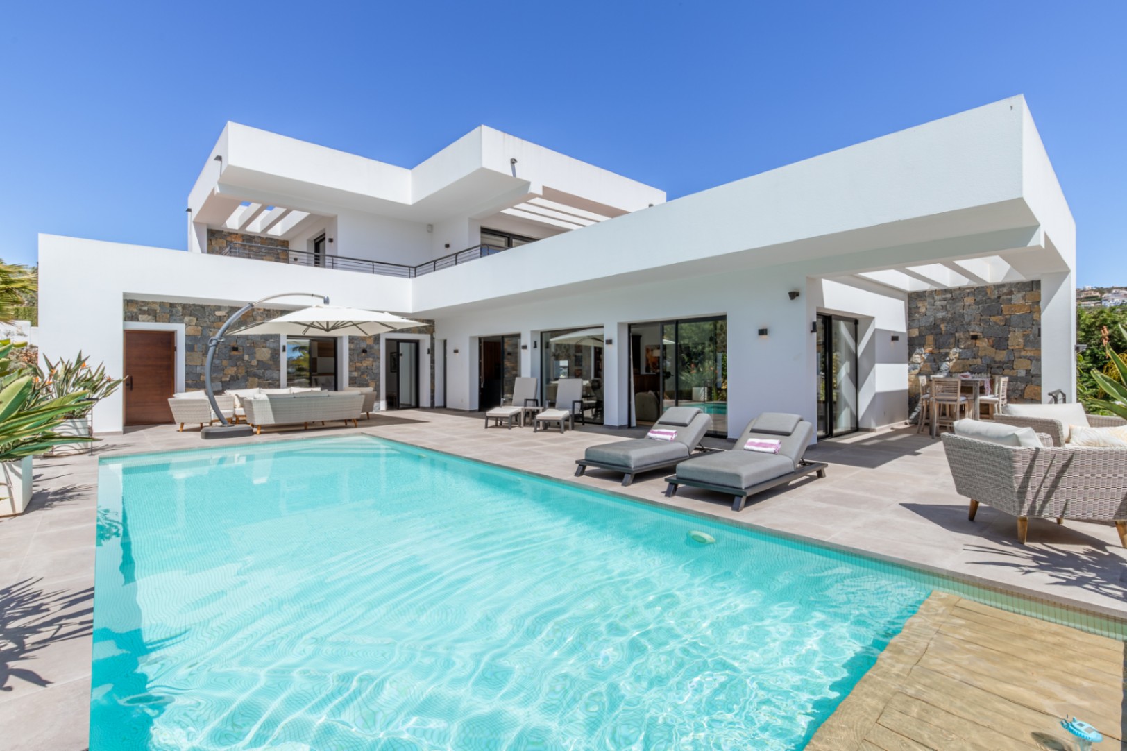 Exterior de la villa con piscina, amplia terraza de gres y fachada blanca con detalles en piedra natural bajo un diseño arquitectónico moderno.