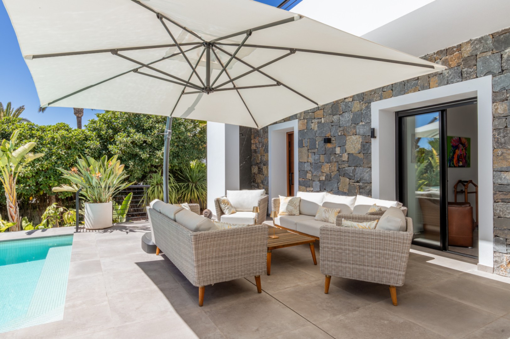 Terraza exterior con zona de estar de mimbre bajo un gran parasol, junto a la piscina y una fachada de piedra natural que aporta textura y privacidad.