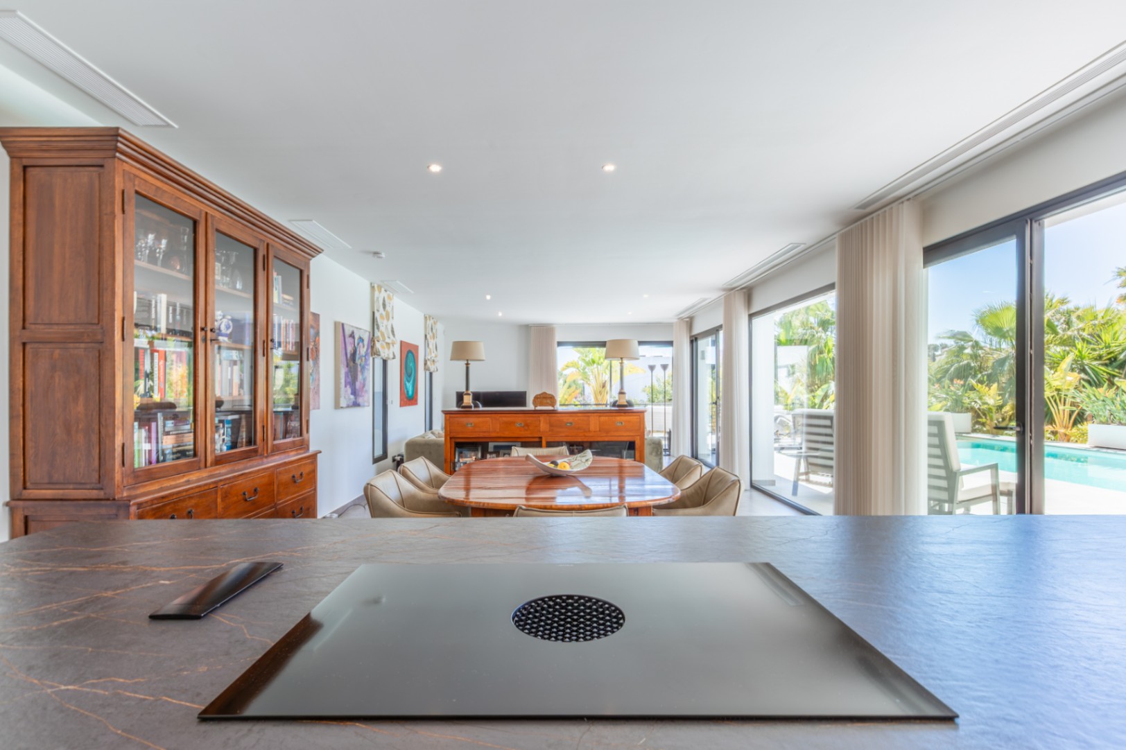 Vista desde la isla de cocina con placa de inducción integrada hacia el comedor, destacando el mobiliario de madera y el acceso directo a la zona de piscina.