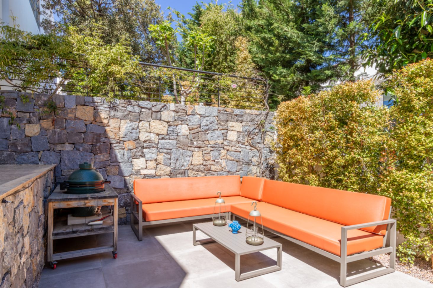 Zona de estar exterior con sofá rinconera naranja, barbacoa cerámica tipo kamado y muro de piedra natural rodeado de vegetación madura.