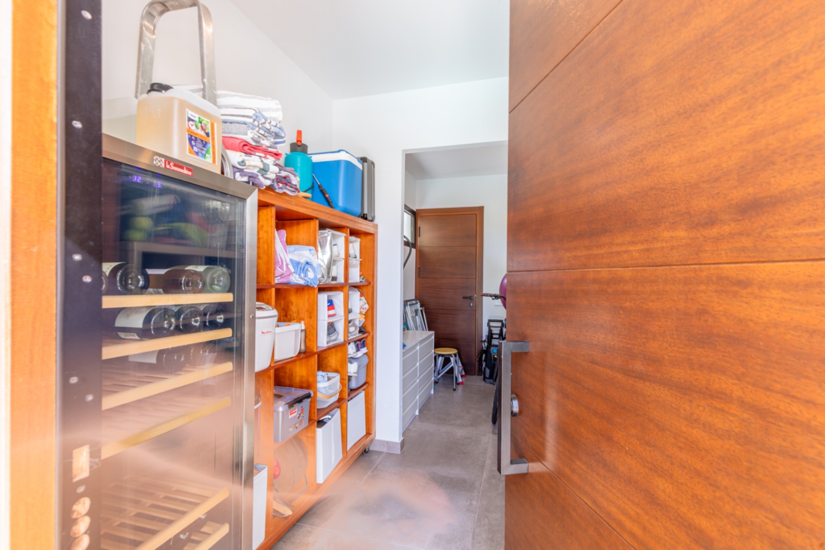 Zona de lavandería y despensa con estanterías de madera, vinoteca integrada y acceso directo, destacando por su funcionalidad y suelos de gres.