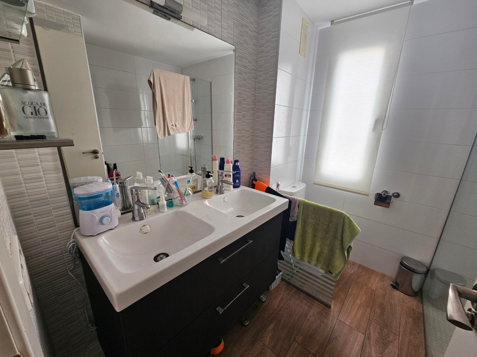 Baño moderno con mueble de doble lavabo en tonos oscuros, suelo de gres cerámico efecto madera y paredes alicatadas en tonos neutros con luz natural.