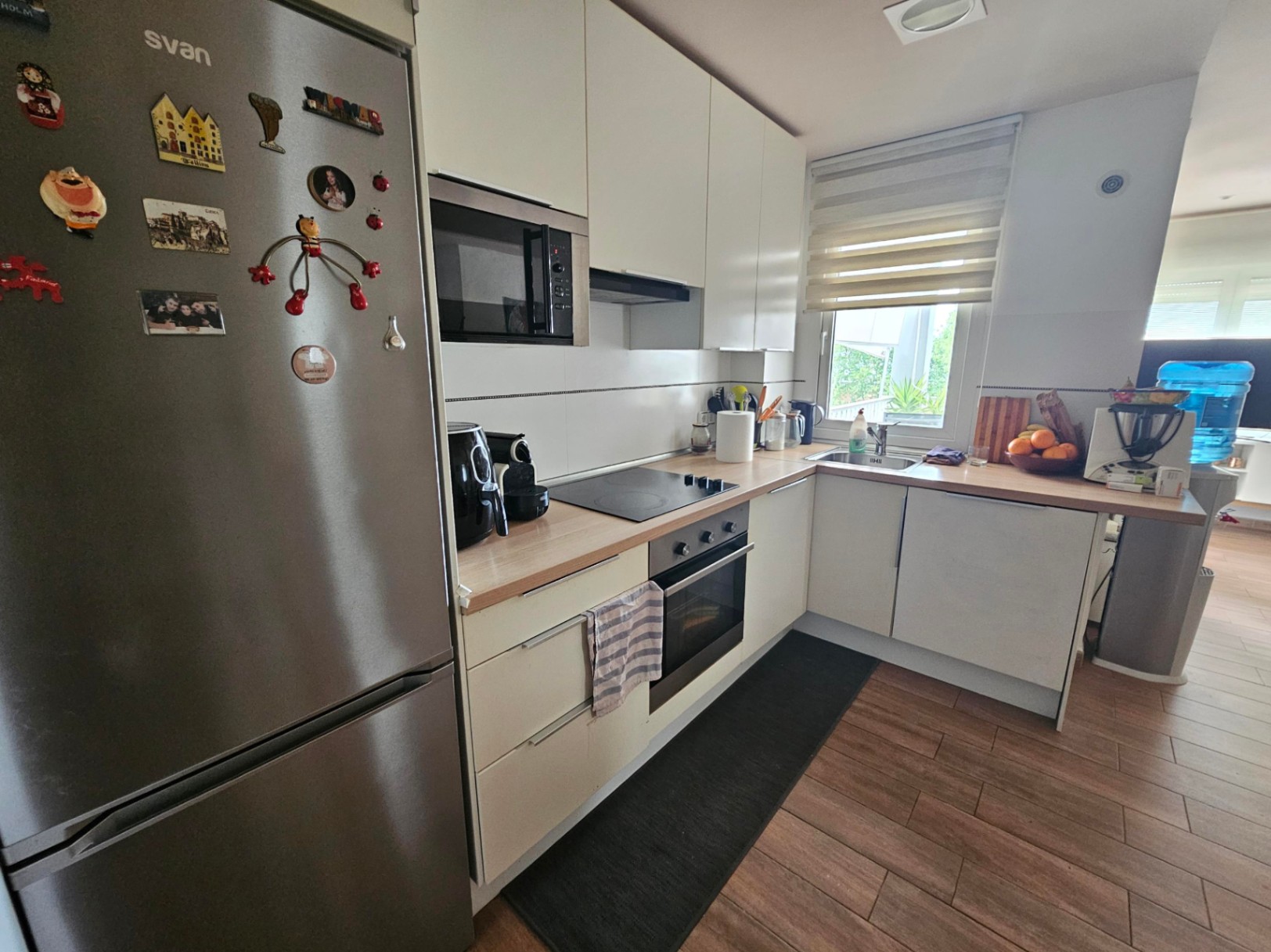 Cocina funcional en forma de L con encimeras de madera, electrodomésticos integrados de acero inoxidable y suelos de gres con efecto madera natural.
