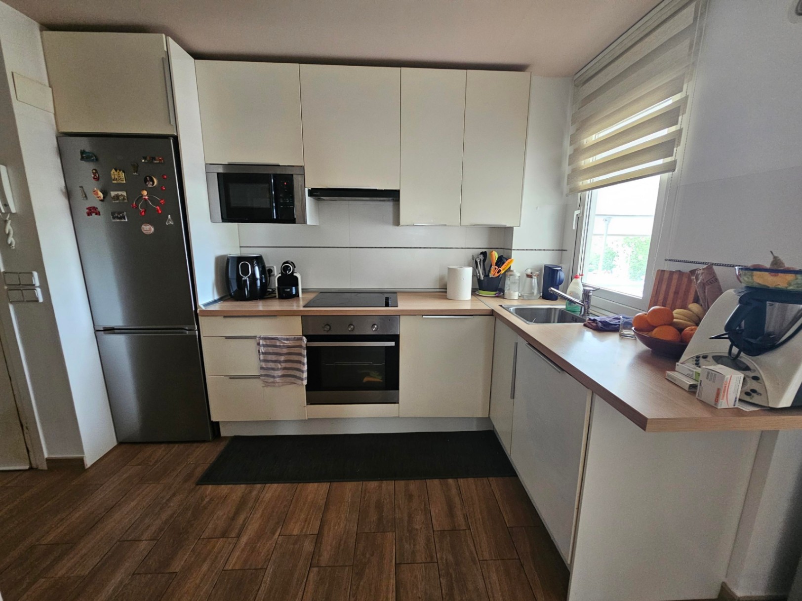 Cocina moderna en forma de L con armarios blancos, encimera de madera, electrodomésticos integrados y suelo cerámico de efecto madera.