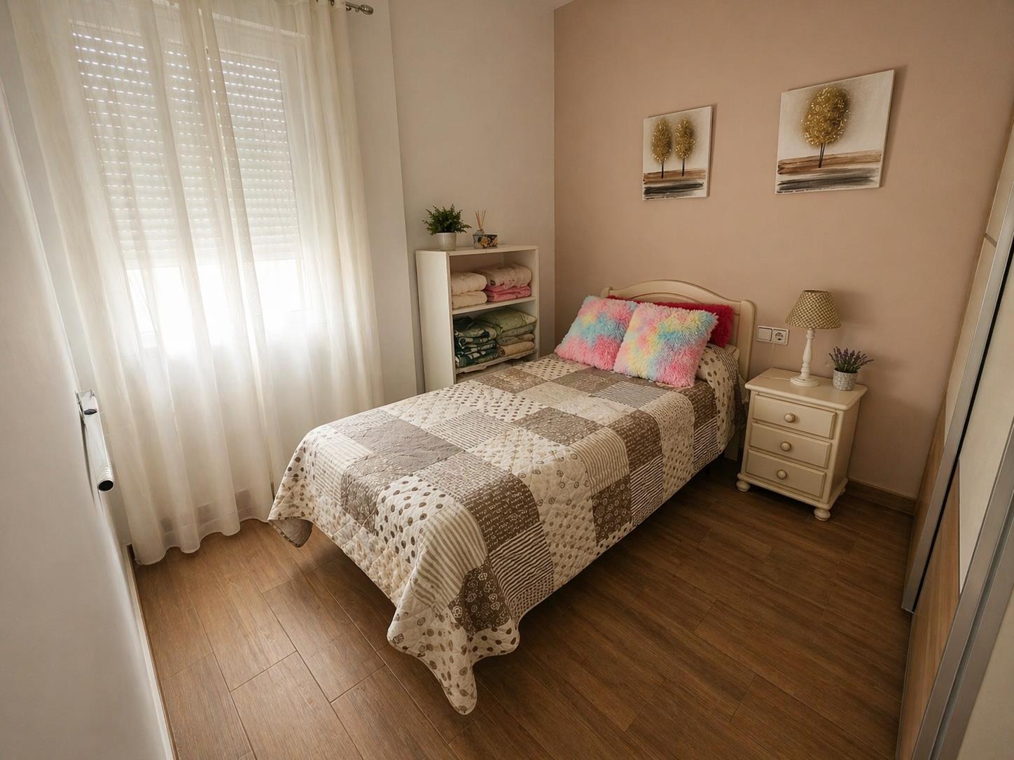 Dormitorio individual con suelos de gres efecto madera, ventana con persiana y cortinas blancas, y una pared de acento en tono salmón con cuadros decorativos.
