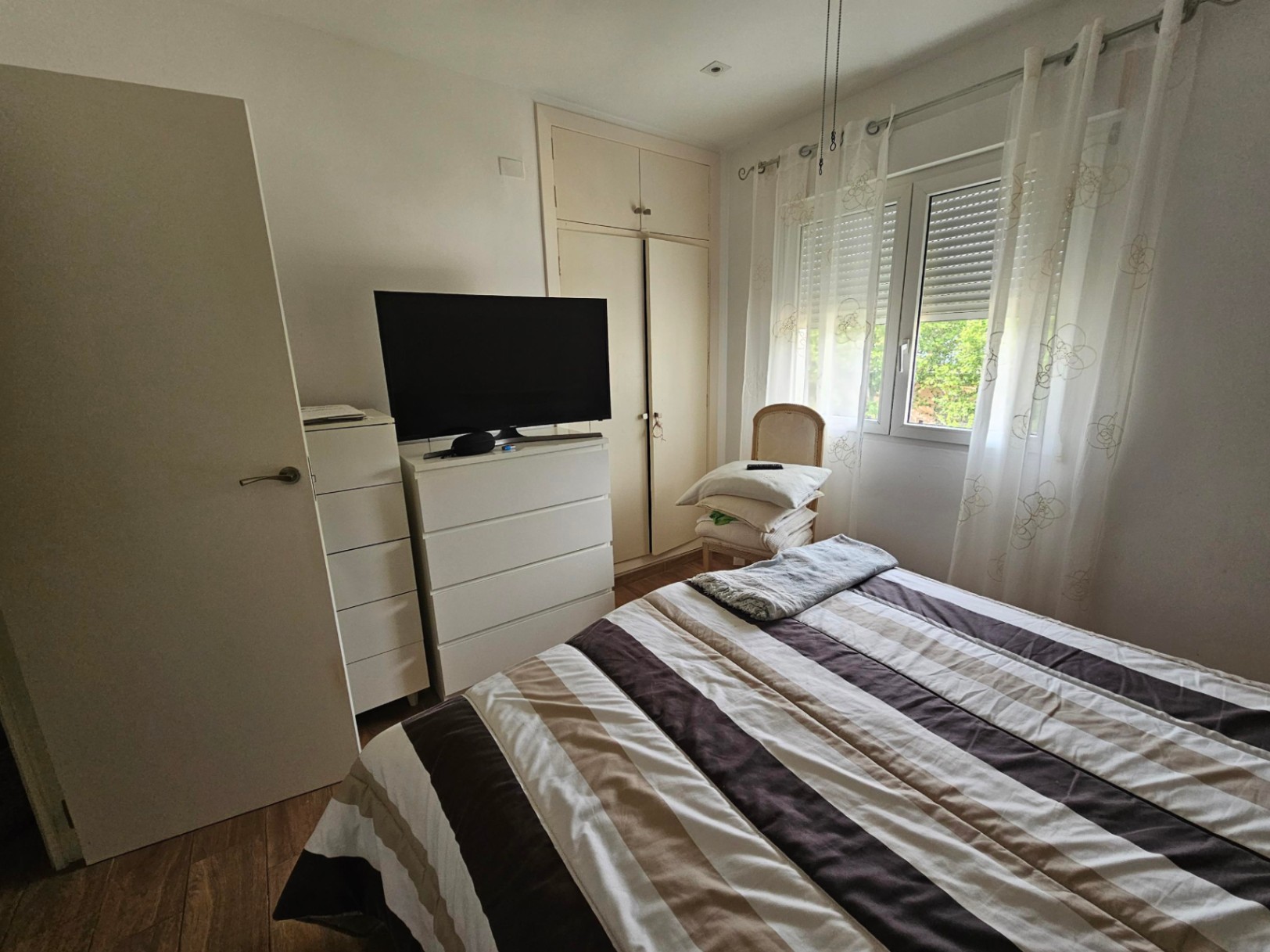 Dormitorio luminoso con cama doble, armarios empotrados, cómoda blanca con televisor y ventana con persianas exteriores y cortinas ligeras.