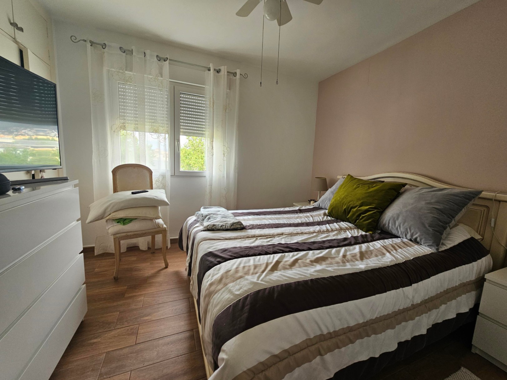 Dormitorio luminoso con pared de acento en tono rosa, suelos de gres efecto madera, ventilador de techo y ventana con persiana y cortinas traslúcidas.