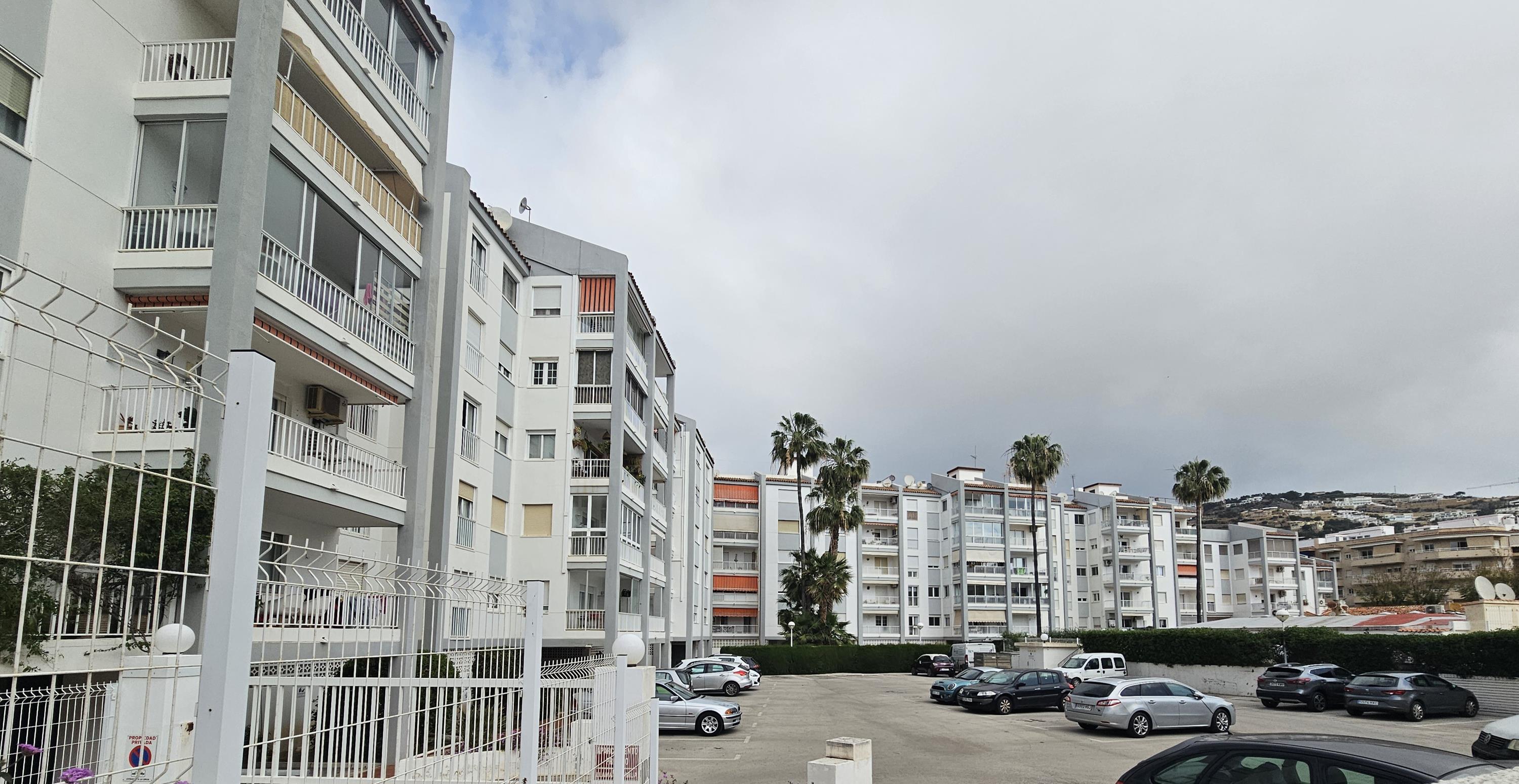 Fachada del edificio residencial con balcones y toldos, que incluye un amplio parking comunitario cerrado y zonas ajardinadas con palmeras.