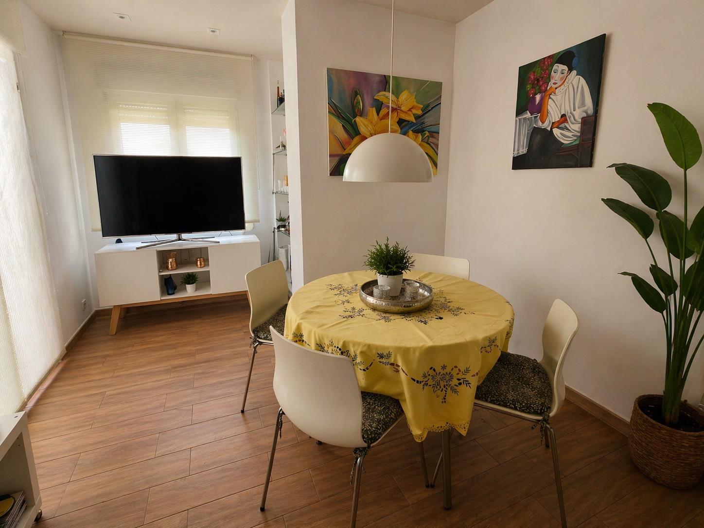 Zona de comedor con mesa redonda, sillas blancas y lámpara de suspensión, integrada en un salón con suelos de efecto madera y luz natural.