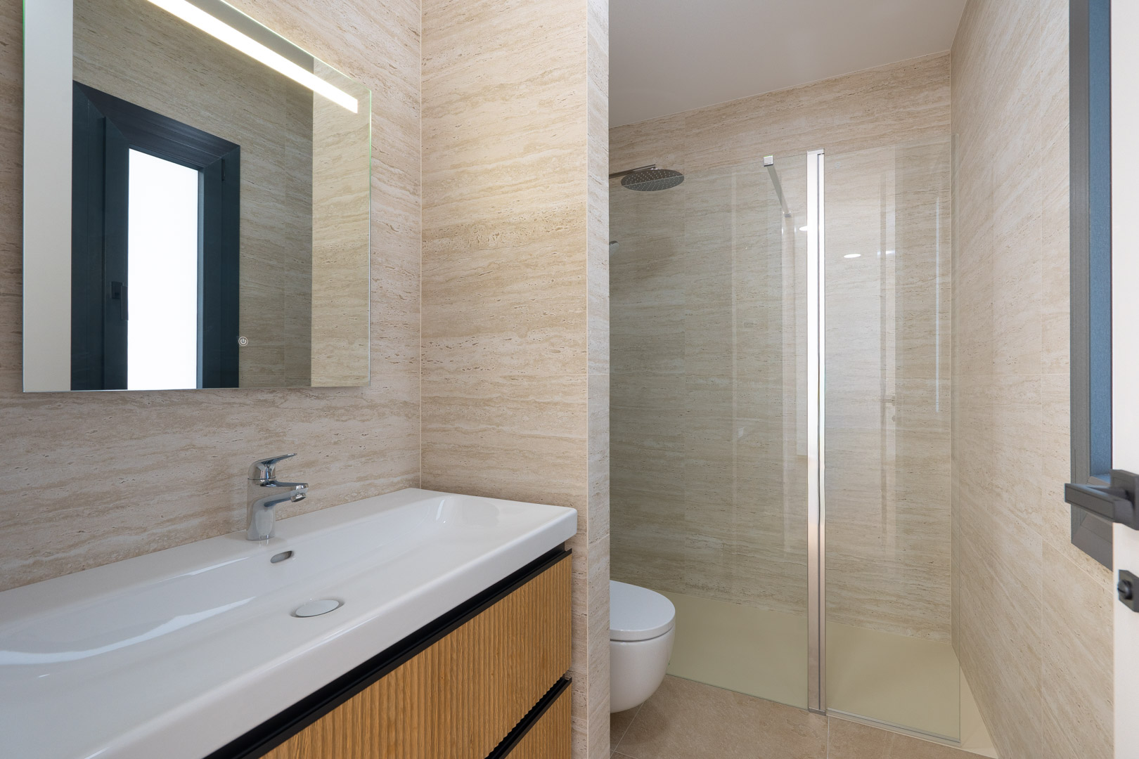 Baño de diseño con revestimiento porcelánico de efecto piedra, lavabo suspendido con mueble de madera estriada y ducha italiana con mampara de cristal y grifería empotrada.