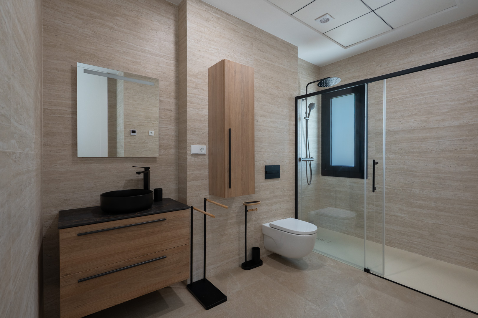 Baño de diseño con revestimiento porcelánico efecto travertino, mueble de madera con lavabo sobre encimera negro y ducha italiana con perfilería negra mate.