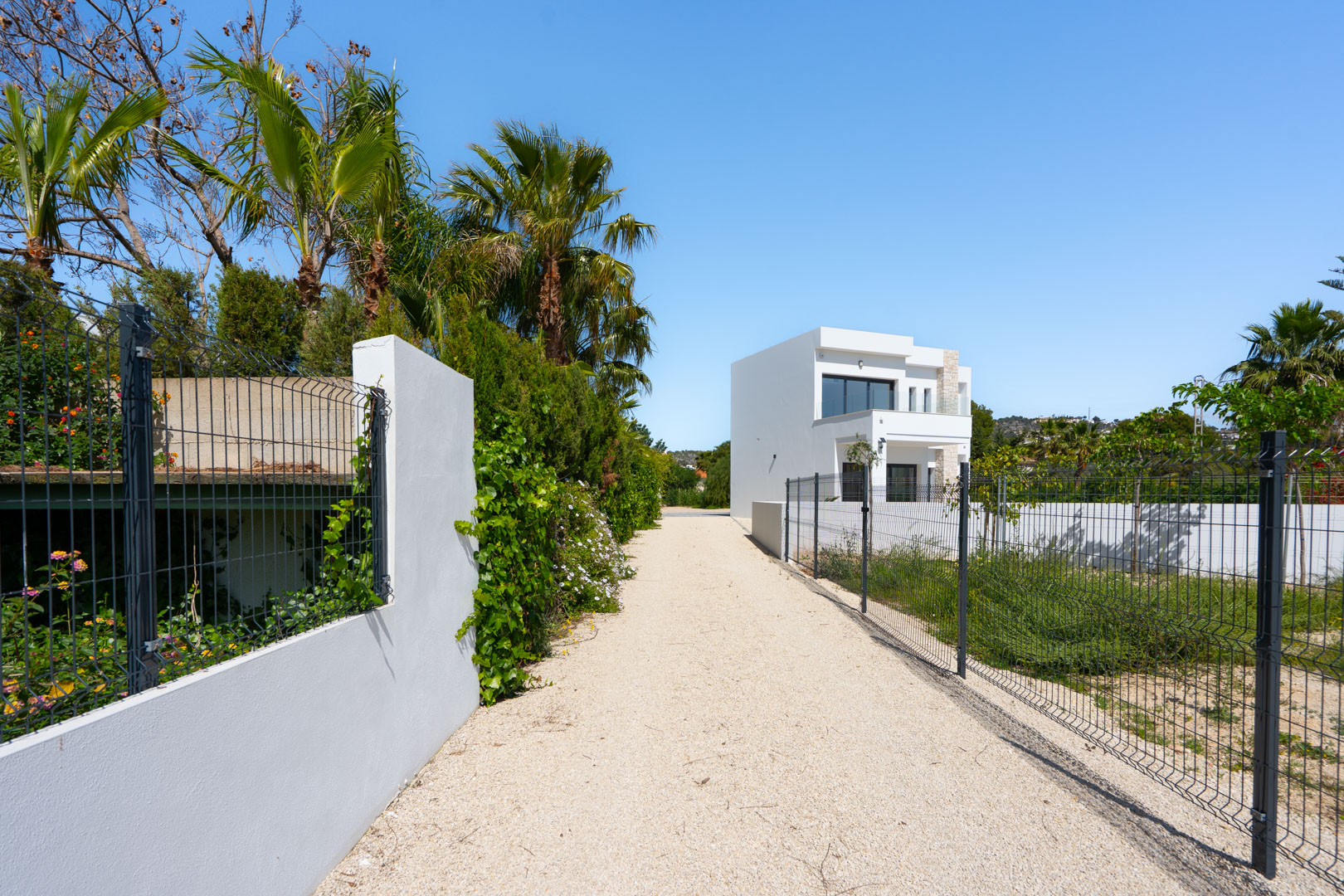 Camino de acceso de grava blanca hacia la villa moderna, flanqueado por muros perimetrales, vegetación mediterránea y una amplia parcela llana vallada.