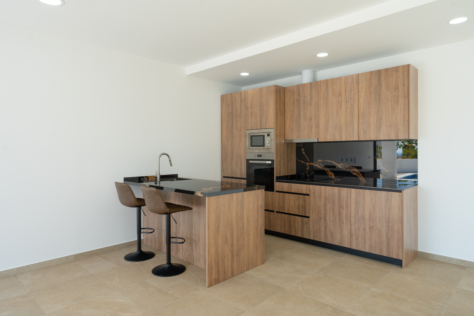 Cocina de diseño con mobiliario en acabado madera, encimera de piedra negra veteada y una isla funcional con taburetes. Incluye electrodomésticos integrados y suelos porcelánicos.