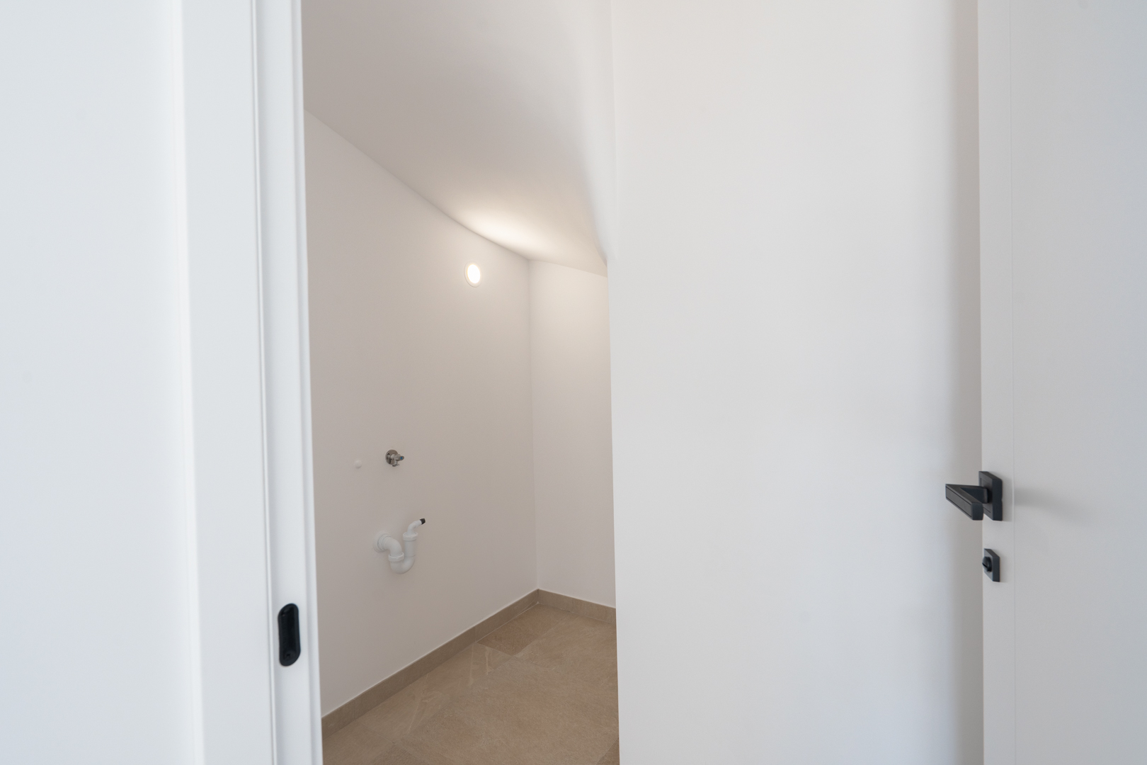 Cuarto de lavandería o trastero con paredes blancas, suelo porcelánico y tomas de agua preparadas bajo el hueco de la escalera.