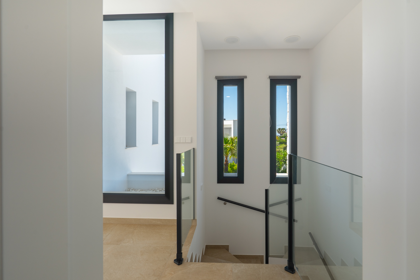 Distribuidor de planta superior con barandillas de cristal, suelos porcelánicos y grandes ventanales que integran luz natural y vistas al jardín mediterráneo.