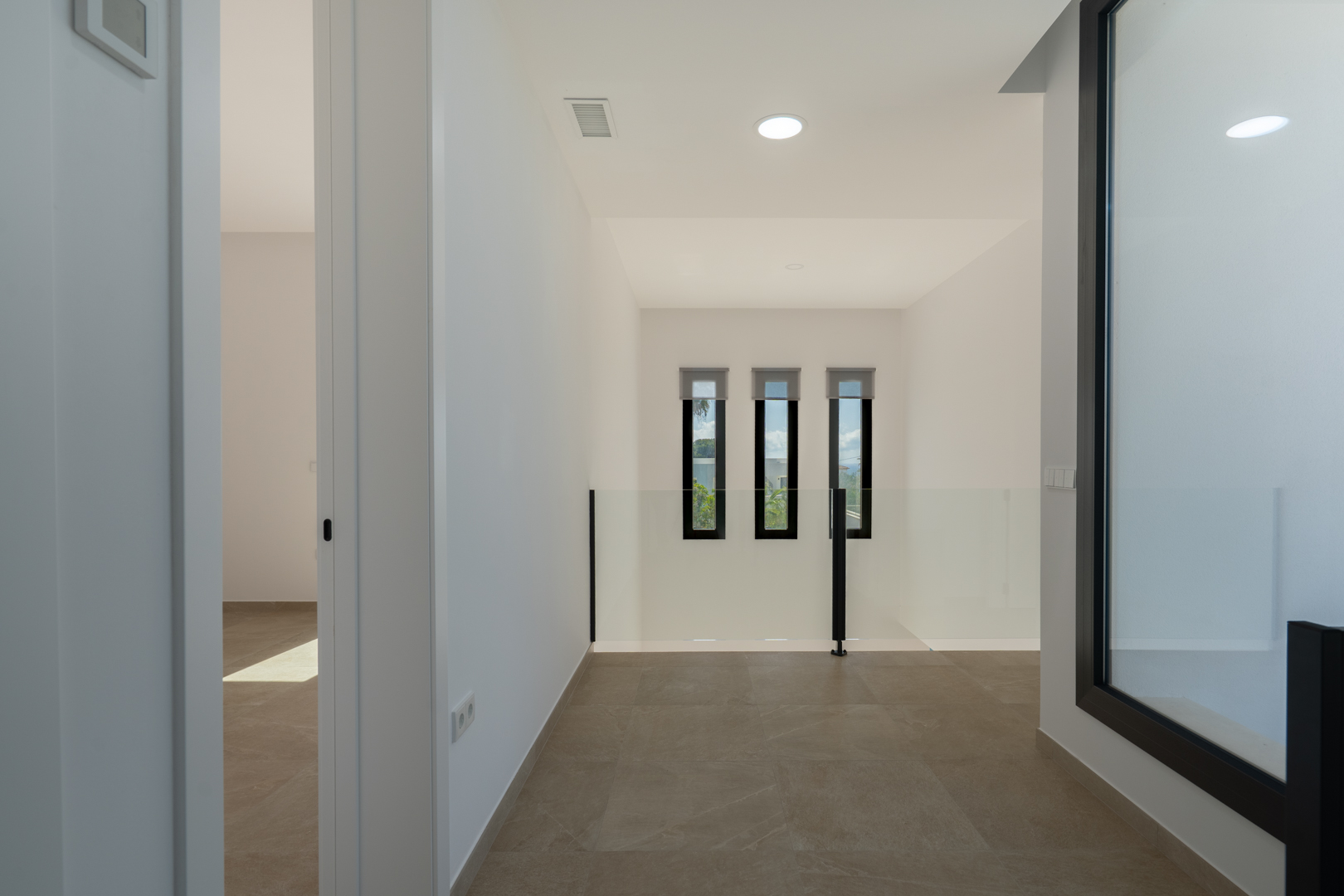 Distribuidor en planta alta con suelos porcelánicos, barandilla de cristal y tres ventanas verticales que aportan luz natural al espacio de doble altura.