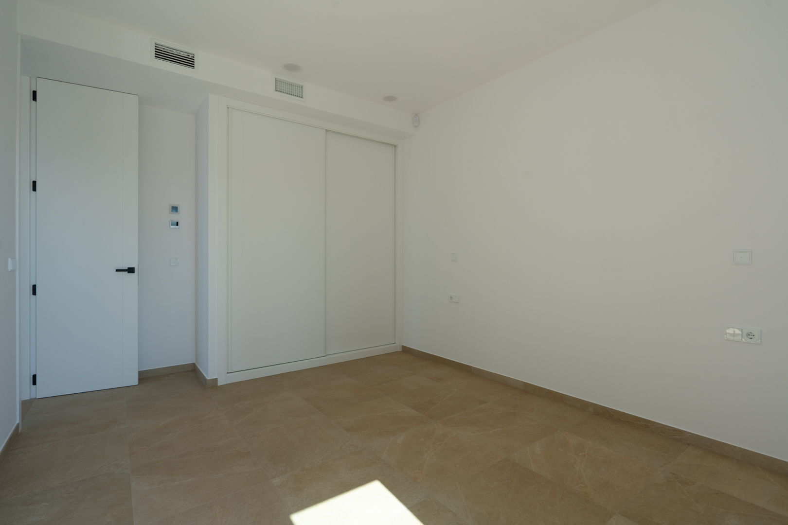 Dormitorio luminoso con suelos porcelánicos en tonos tierra, armario empotrado de puertas correderas blancas y sistema de climatización por conductos integrado.