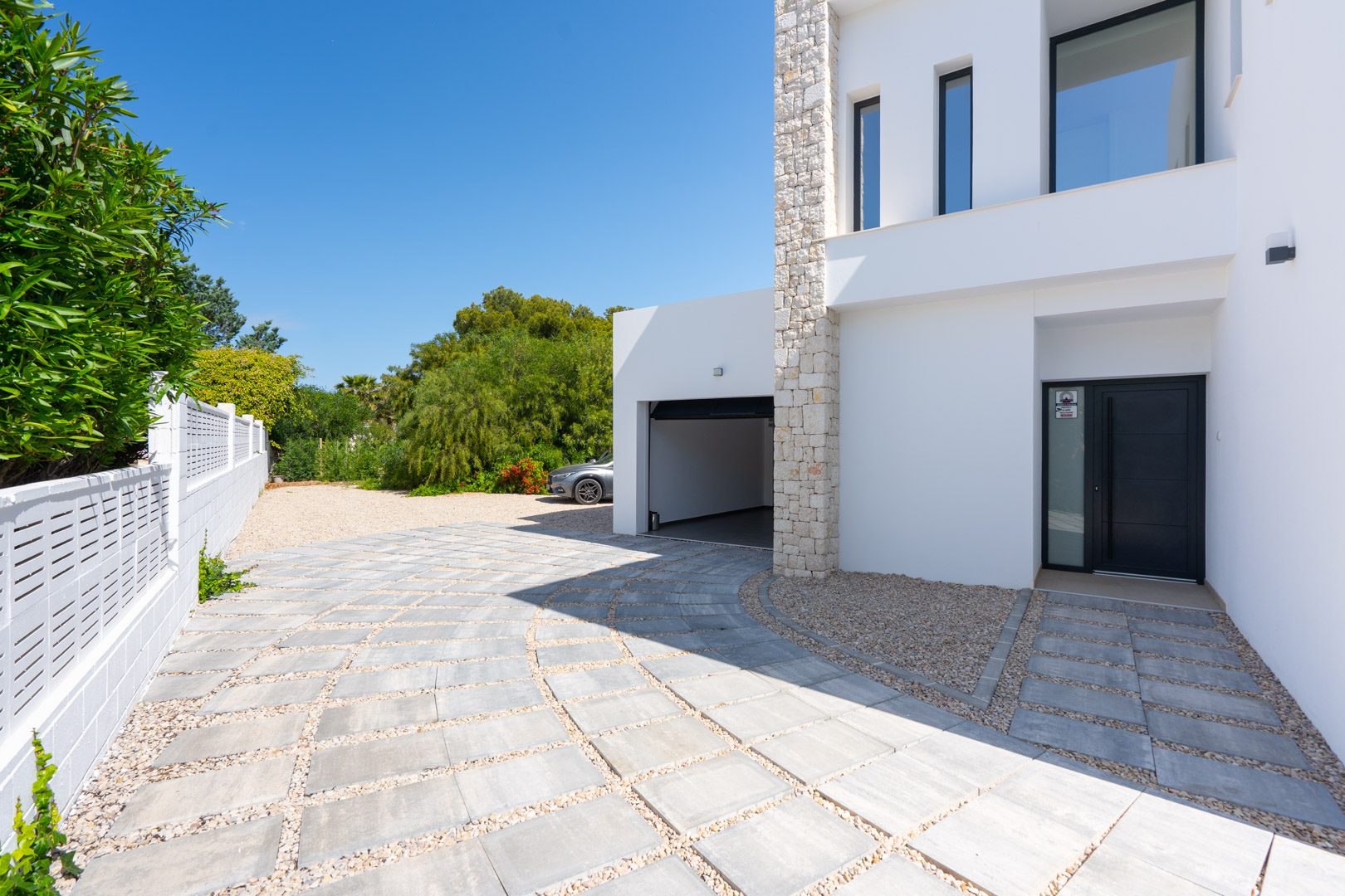 Entrada principal y acceso al garaje con pavimentación de baldosas y grava, destacando la fachada blanca con muro decorativo de piedra natural.