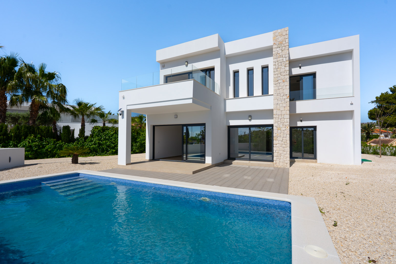 Fachada moderna de dos niveles con muro de piedra natural, piscina de gresite azul y terrazas con barandillas de cristal en un entorno ajardinado con palmeras.