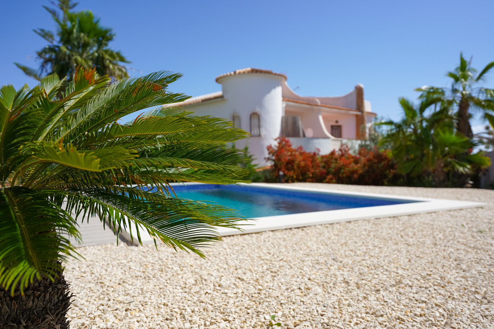Piscina exterior rodeada de grava blanca y vegetación mediterránea, con la arquitectura de la villa y su torre característica al fondo bajo el sol.
