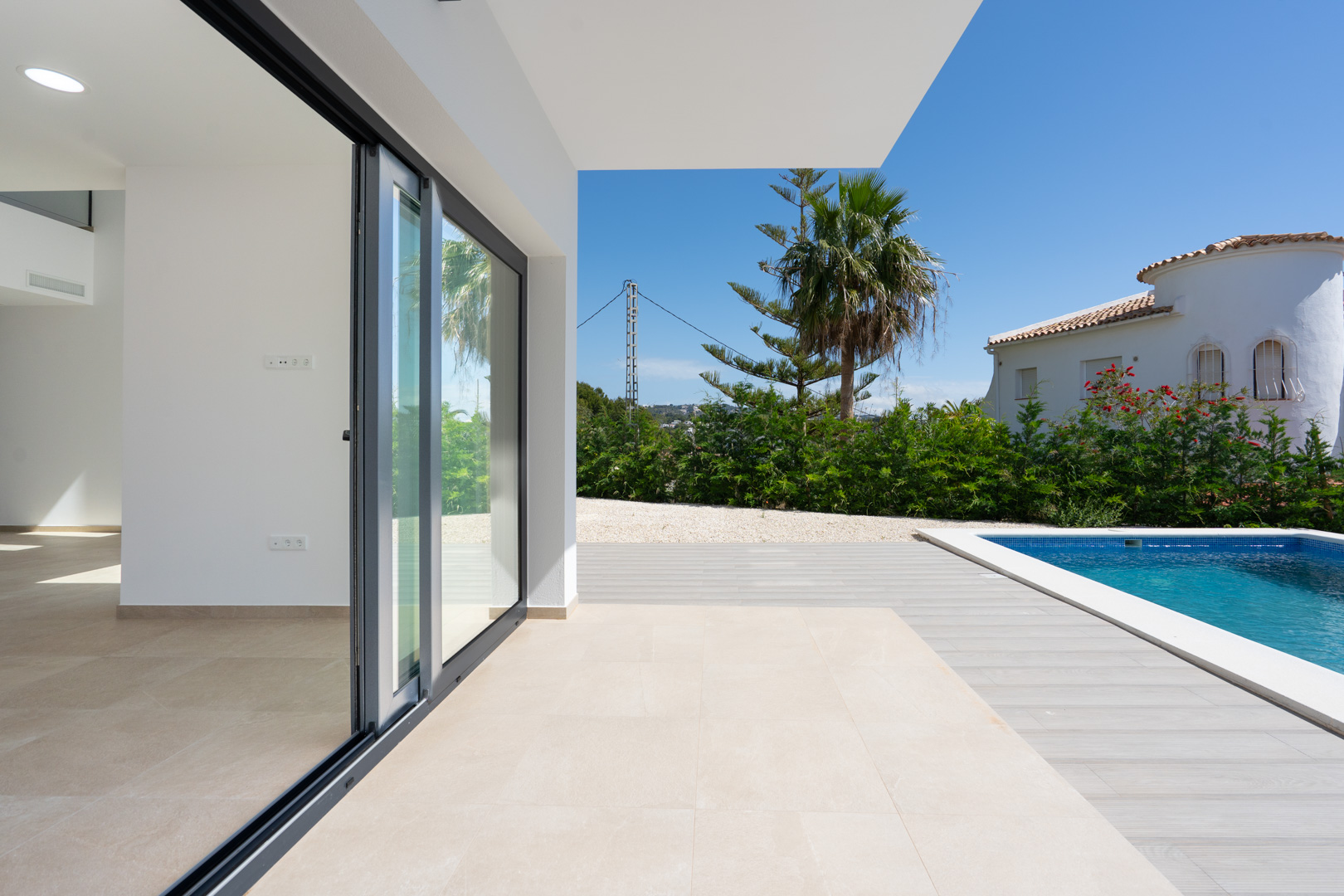 Terraza cubierta con suelos porcelánicos que conectan el interior con la piscina de mosaico azul, rodeada de vegetación mediterránea y palmeras.