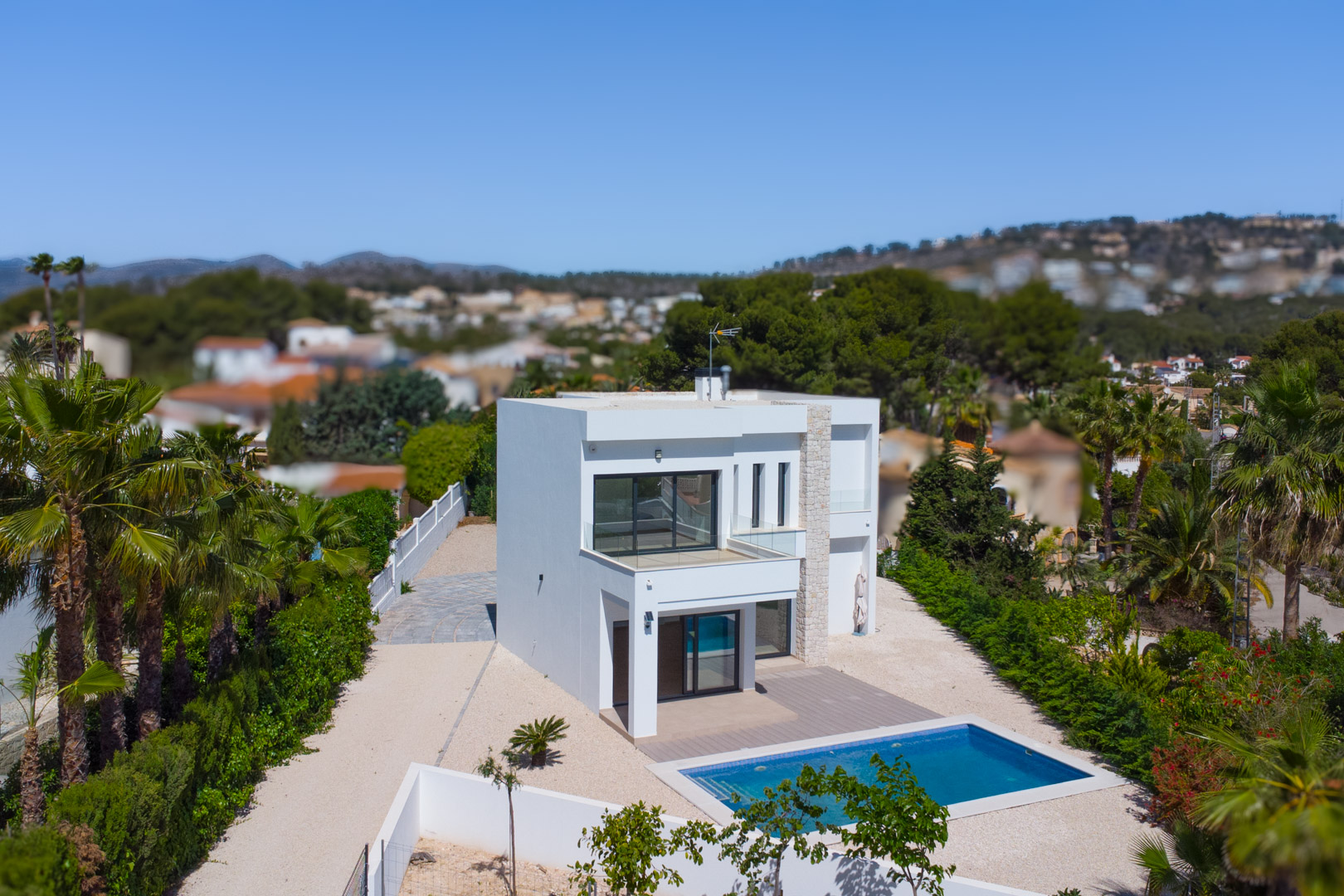 Vista exterior de la villa moderna con fachada blanca, detalles en piedra natural, piscina rectangular y terrazas con barandillas de cristal sobre una gran parcela llana.