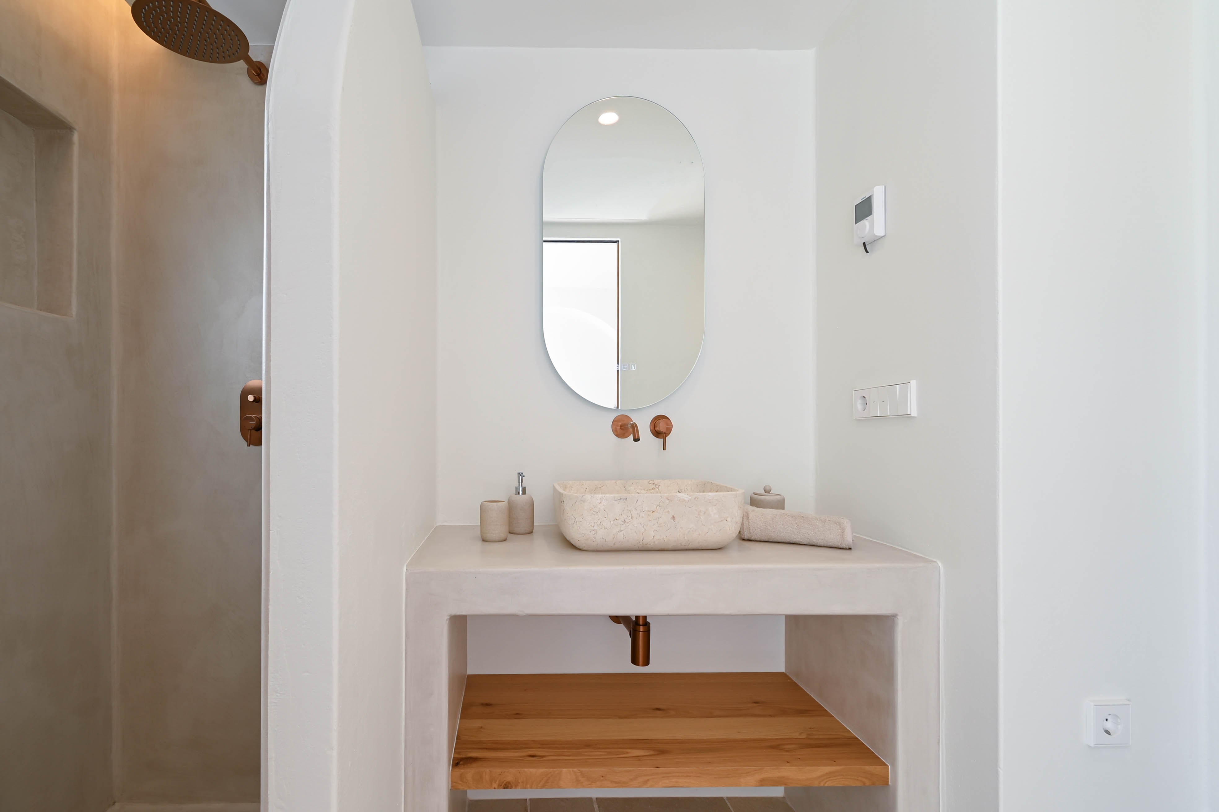 Baño moderno con acabados de microcemento, lavabo de piedra natural y grifería en tono cobre. Incluye un estante inferior de madera y ducha de diseño minimalista.