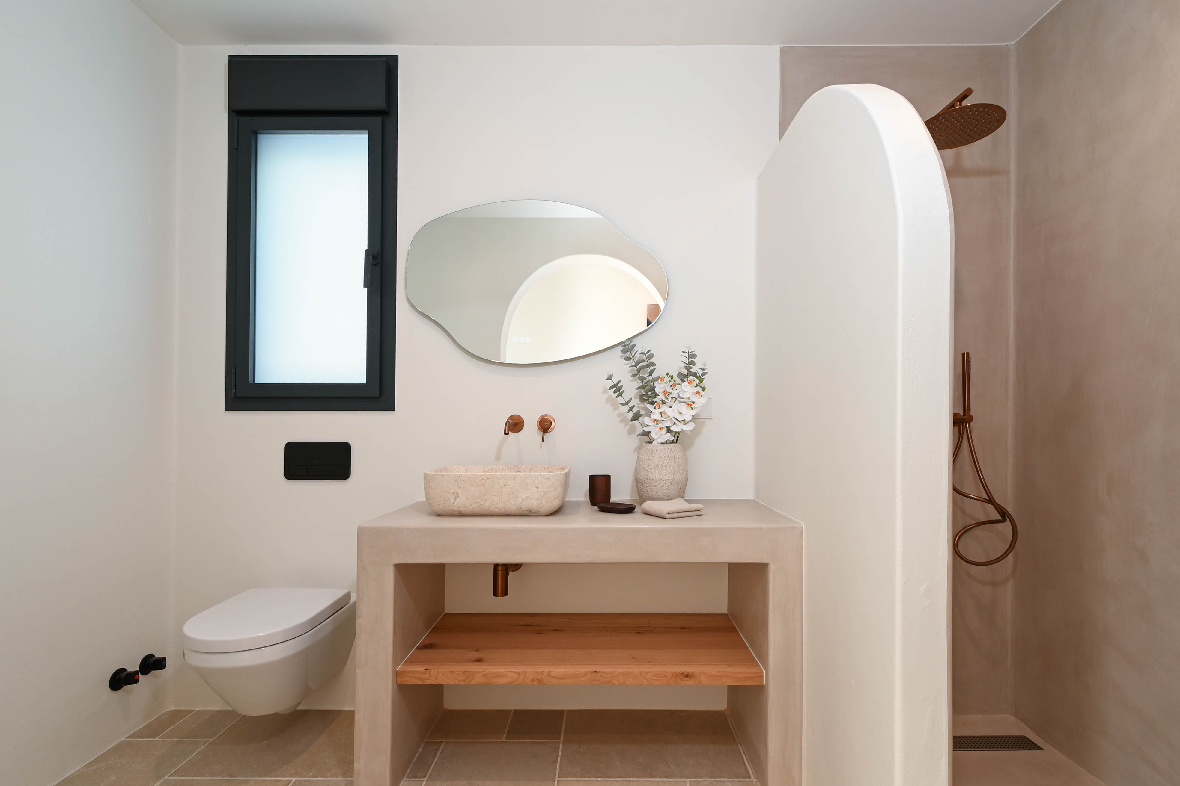 Baño moderno con encimera de microcemento, lavabo de piedra natural y grifería de cobre. Incluye un espejo orgánico, inodoro suspendido y ducha de obra con pared divisoria curva.