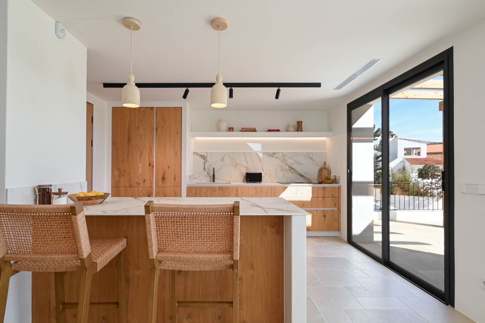 Cocina de concepto abierto con isla de madera, encimera de efecto mármol y taburetes de mimbre, conectada a la terraza exterior a través de amplios ventanales negros.
